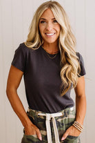 A smiling blonde woman wearing a dark charcoal gray short-sleeve t-shirt and camouflage pants with a white drawstring, standing against a light paneled wall.