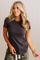 A woman wearing a dark charcoal gray short-sleeve crewneck t-shirt and camouflage pants, standing against a white paneled wall.