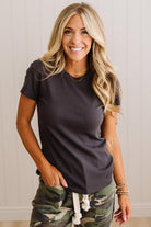 A smiling blonde woman wearing a dark charcoal gray short-sleeve crewneck t-shirt and camouflage pants, standing against a light beige paneled wall.