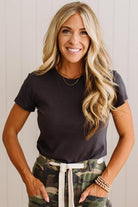 A smiling blonde woman wearing a dark charcoal gray short-sleeve crewneck t-shirt and camouflage pants with a white drawstring, standing against a light beige paneled wall.