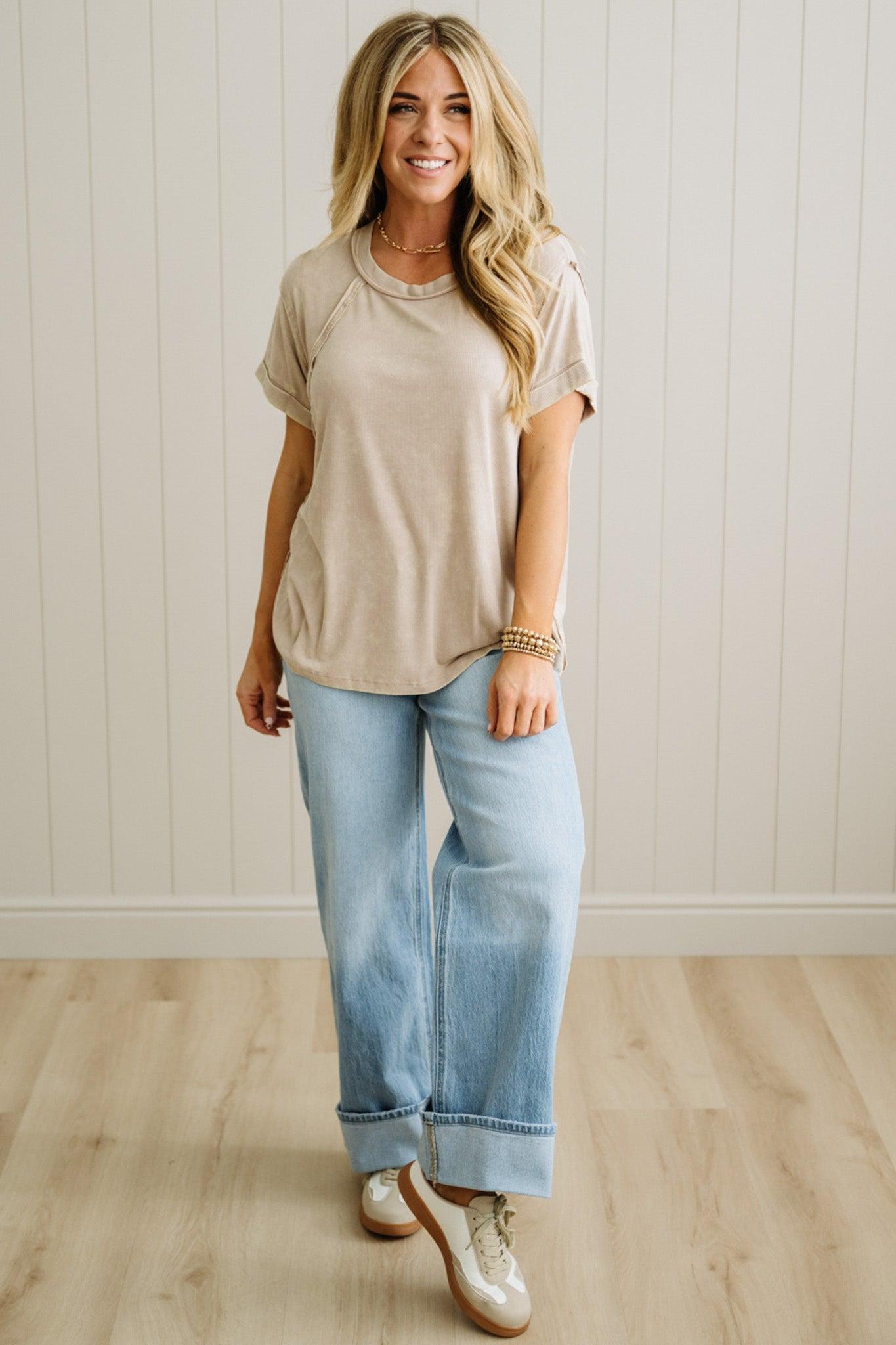 Woman wearing a beige t-shirt and light blue jeans standing against a white paneled wall.