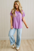 Woman wearing a purple t-shirt and light blue jeans standing against a white paneled wall.