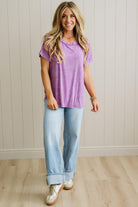 Woman wearing a purple top and light blue jeans standing against a white paneled wall.