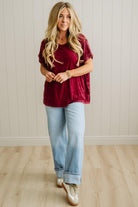 Woman wearing a burgundy top and light blue jeans standing against a white paneled wall.