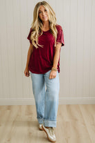 Woman wearing a burgundy top and light blue jeans standing against a white paneled wall.