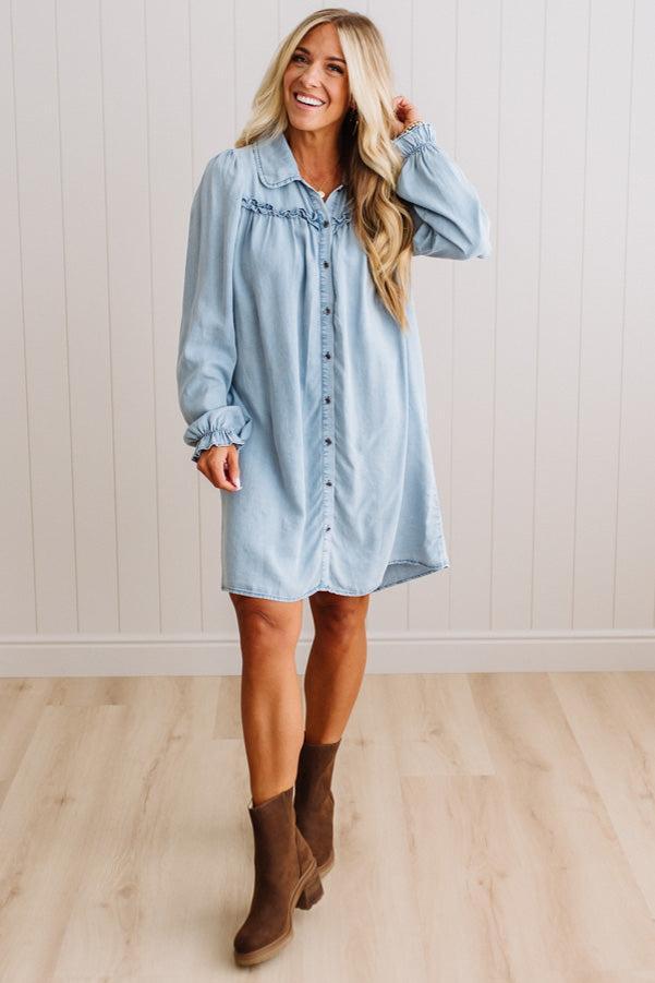 A woman modeling a light blue denim button-up shirt dress with ruffle details on the chest and cuffs, paired with brown ankle boots, standing in front of a white paneled wall and light wood floor.