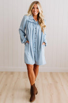 A woman modeling a light blue denim shirt dress with ruffled trim at the collar and button-front closure, paired with brown ankle boots, standing against a white paneled wall and light wood floor.