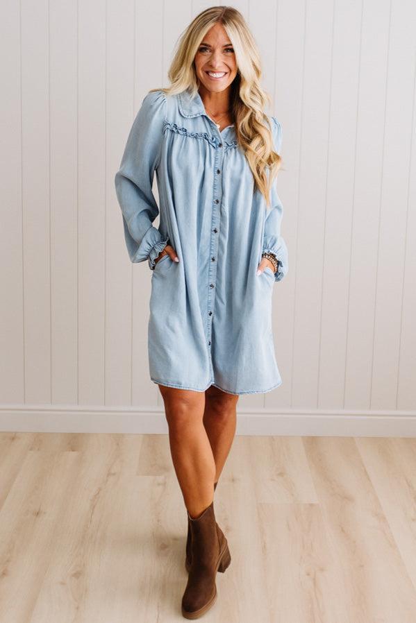 A woman modeling a light blue denim shirt dress with ruffled trim at the collar and button-front closure, paired with brown ankle boots, standing against a white paneled wall and light wood floor.