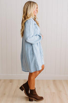 A woman modeling a light blue denim shirt dress with brown ankle boots, standing in profile against a white paneled wall and light wood floor.