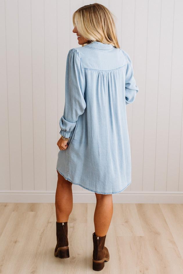 A woman wearing a light blue denim shirt dress, shown from the back. She is standing on a light wood floor against a white paneled wall.