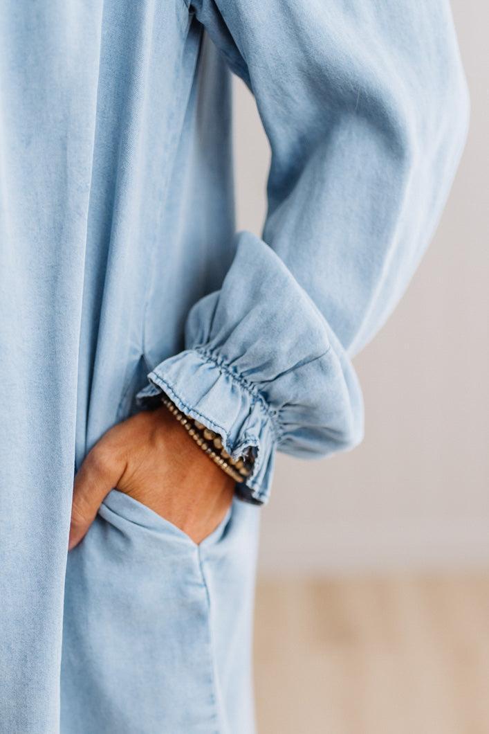 Close-up of a person wearing a light blue long-sleeve top or dress, with one hand tucked into a front pocket and the other near the gathered cuff.