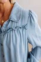Close-up of a person wearing a light blue denim shirt with ruffled trim across the chest and a collar, against a soft neutral background.