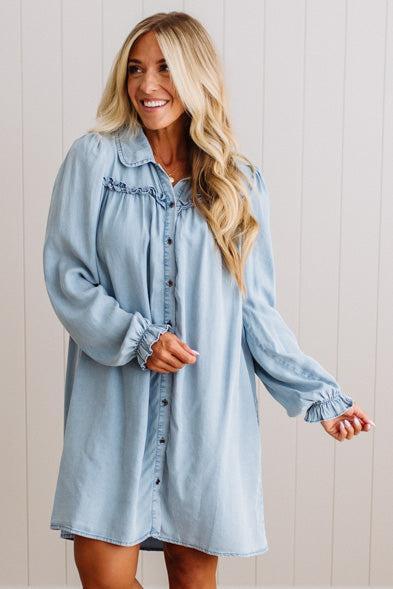 A woman wearing a light blue denim button-up shirt dress with ruffled details on the chest and cuffs, standing against a white paneled wall.