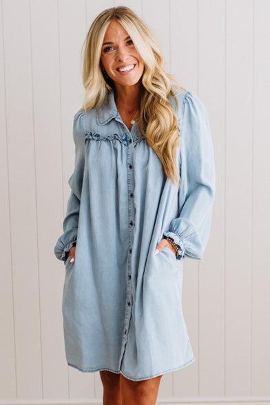 A woman wearing a light blue denim shirt dress with ruffled trim at the collar and button-front pockets, standing against a white paneled wall.