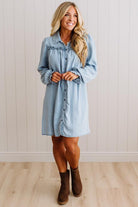A woman modeling a light blue denim shirt dress with ruffle details on the chest and cuffs, paired with brown ankle boots, standing against a white paneled wall and light wood floor.