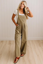 A woman modeling a pair of wide-leg olive green jumpsuit overalls over a white t-shirt and brown sandals, standing in a room with white paneled walls and light wood floors.