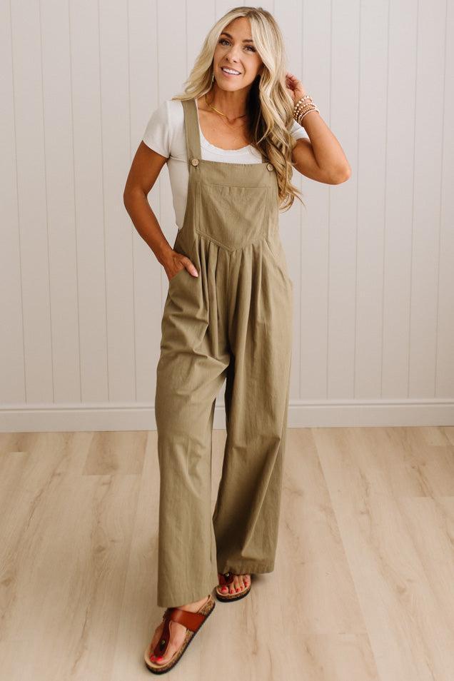 A woman modeling a pair of wide-leg olive green jumpsuit overalls over a white t-shirt and brown sandals, standing in a room with white paneled walls and light wood floors.