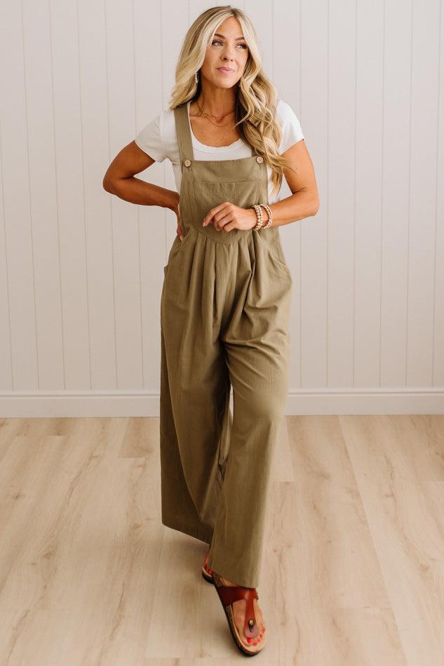 A woman modeling a pair of olive green wide-leg utility jumpsuits over a white t-shirt, standing on a light wood floor against a white paneled wall.