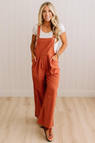 A woman modeling a rust-orange wide-leg overall jumpsuit over a white t-shirt, standing in a room with light wood floors and white paneled walls.