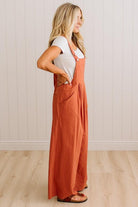A woman wearing a rust-orange linen wide-leg jumpsuit over a white t-shirt, standing in profile against a white paneled wall and light wood floor.