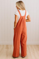 A woman viewed from behind wearing rust-orange wide-leg overall jumpsuit over a white t-shirt, standing on a light wood floor against a white paneled wall.