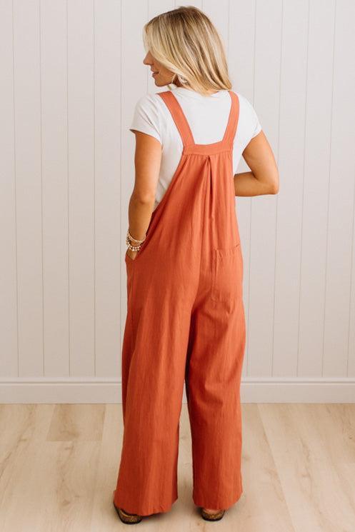 A woman viewed from behind wearing rust-orange wide-leg overall jumpsuit over a white t-shirt, standing on a light wood floor against a white paneled wall.