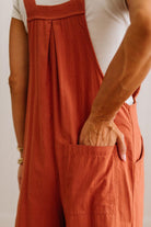 A close-up of a person wearing rust-orange bib overalls with a large front patch pocket, shown from the chest down against a plain light background.