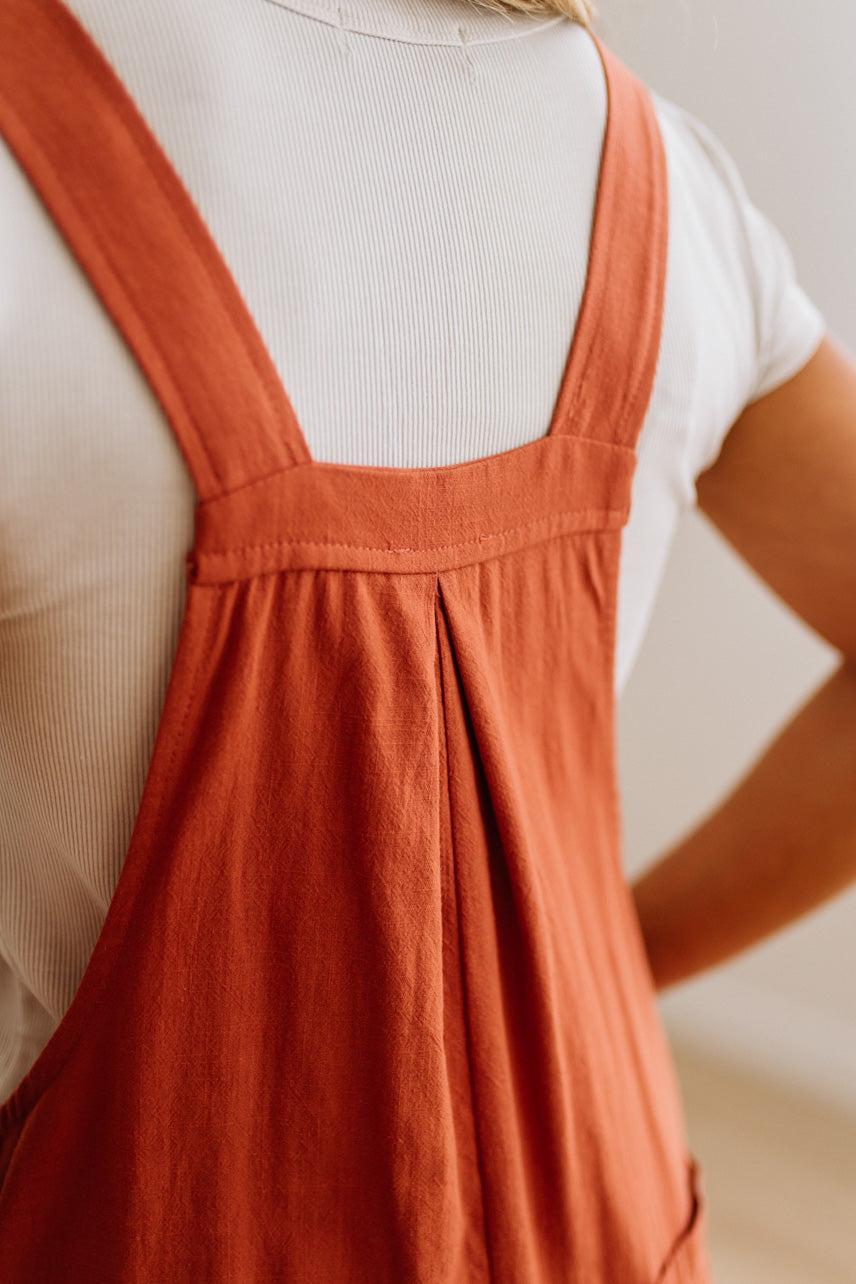 Close-up of a person wearing rust-orange overall-style shorts over a white ribbed T-shirt, with the focus on the garment's back and shoulder straps.