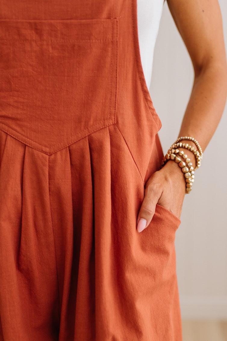 Close-up of a terracotta rust-colored linen overall jumpsuit with a front pocket and pleated skirt detail, worn by a model with a hand in the pocket and gold beaded bracelets.