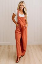 A woman modeling a rust-orange linen overall jumpsuit with a white t-shirt underneath, standing on a light wood floor against a white paneled wall.