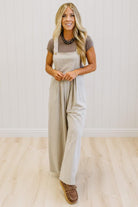 A woman modeling a light beige linen overall jumpsuit over a taupe short-sleeve t-shirt, standing on a light wood floor against a white paneled wall.