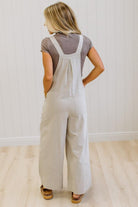 A woman modeling a pair of wide-leg stone colored utility jumpsuit over a brown t-shirt, standing in a room with light wood floors and white paneled walls.