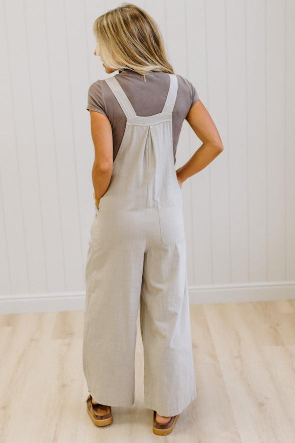 A woman modeling a pair of wide-leg stone colored utility jumpsuit over a brown t-shirt, standing in a room with light wood floors and white paneled walls.