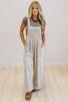 A woman modeling a pair of wide-leg stone colored utility jumpsuit over a brown t-shirt, standing in a room with light wood floors and white paneled walls.