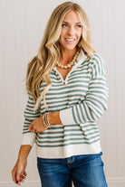 BLONDE WOMAN IN DARK JEANS STANDING IN A BRIGHTLY LIT STUDIO WITH LIGHT WOOD FLOORS AND A PANELED BACKDROP. SHE IS WEARING A CHUNKY NECKLACE AND QUARTER ZIP STRIPES SWEATER IN CREAM AND GREEN.