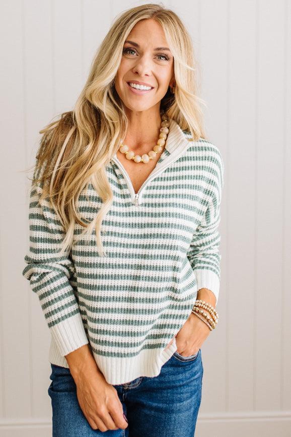 BLONDE WOMAN IN DARK JEANS STANDING IN A BRIGHTLY LIT STUDIO WITH LIGHT WOOD FLOORS AND A PANELED BACKDROP. SHE IS WEARING A CHUNKY NECKLACE AND QUARTER ZIP STRIPES SWEATER IN CREAM AND GREEN.