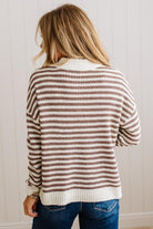 BLONDE WOMAN IN DARK JEANS STANDING IN A BRIGHTLY LIT STUDIO WITH LIGHT WOOD FLOORS AND A PANELED BACKDROP. SHE IS WEARING A CHUNKY NECKLACE AND QUARTER ZIP STRIPES SWEATER IN CREAM AND MOCHA.