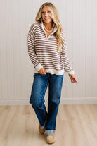 BLONDE WOMAN IN DARK JEANS STANDING IN A BRIGHTLY LIT STUDIO WITH LIGHT WOOD FLOORS AND A PANELED BACKDROP. SHE IS WEARING A CHUNKY NECKLACE AND QUARTER ZIP STRIPES SWEATER IN CREAM AND MOCHA.