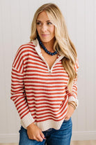 BLONDE WOMAN IN DARK JEANS STANDING IN A BRIGHTLY LIT STUDIO WITH LIGHT WOOD FLOORS AND A PANELED BACKDROP. SHE IS WEARING A CHUNKY NECKLACE AND QUARTER ZIP STRIPES SWEATER IN CREAM AND RUST.