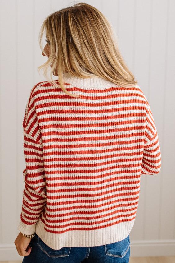 BLONDE WOMAN IN DARK JEANS STANDING IN A BRIGHTLY LIT STUDIO WITH LIGHT WOOD FLOORS AND A PANELED BACKDROP. SHE IS WEARING A CHUNKY NECKLACE AND QUARTER ZIP STRIPES SWEATER IN CREAM AND RUST.