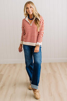 BLONDE WOMAN IN DARK JEANS STANDING IN A BRIGHTLY LIT STUDIO WITH LIGHT WOOD FLOORS AND A PANELED BACKDROP. SHE IS WEARING A CHUNKY NECKLACE AND QUARTER ZIP STRIPES SWEATER IN CREAM AND RUST.