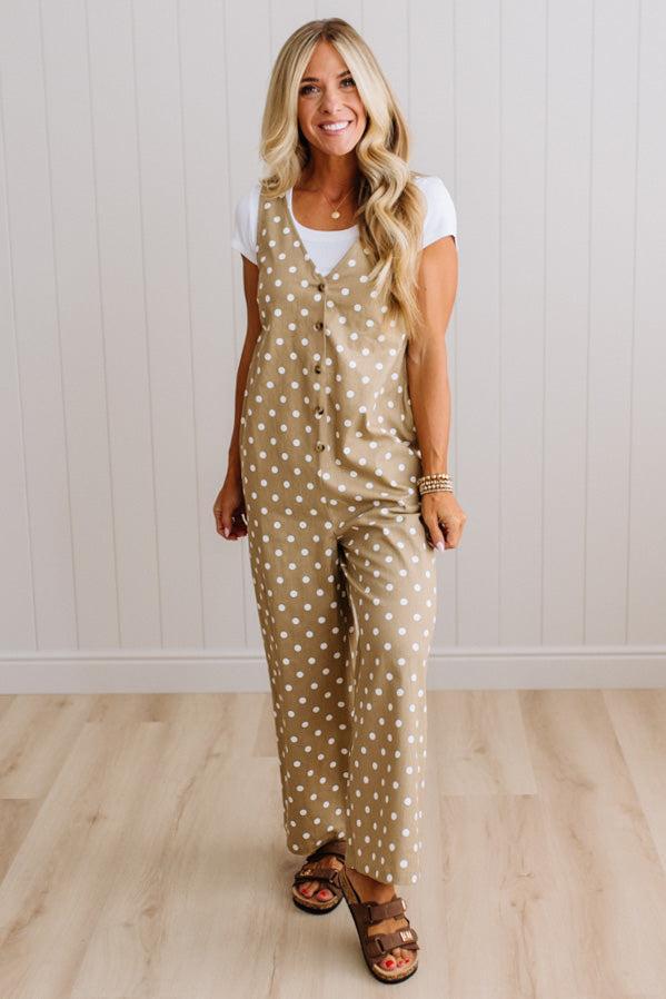 A woman modeling a beige sleeveless jumpsuit with white polka dots, worn over a white t-shirt and brown heeled sandals, standing in front of a white paneled wall and light wood floor.