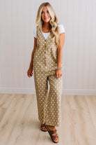 A woman modeling a beige sleeveless jumpsuit with white polka dots, styled over a white t-shirt and brown sandals, standing against a white paneled wall and light wood floor.