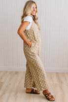 A woman modeling a beige and white polka dot overall jumpsuit with a white t-shirt underneath and brown slide sandals, standing in a room with white paneled walls and light wood floors.