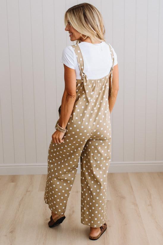 A woman shown from the back wearing beige wide-leg overall jumpsuit with white polka dots over a white t-shirt, standing on a light wood floor against a white paneled wall.