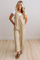 A woman modeling a beige sleeveless jumpsuit with white polka dots over a white t-shirt and brown sandals, standing in front of a white paneled wall and light wood floor.