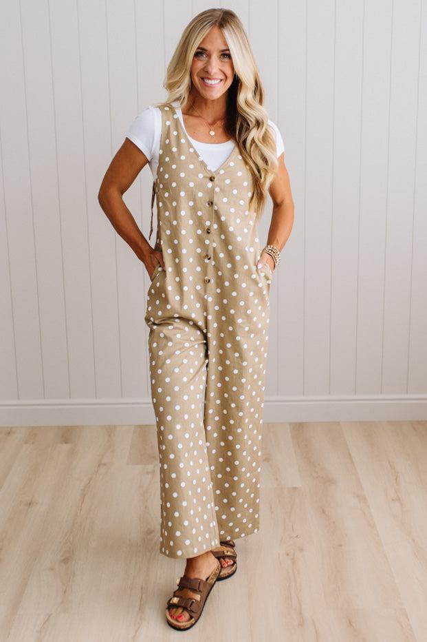 A woman modeling a beige sleeveless jumpsuit with white polka dots over a white t-shirt and brown sandals, standing in front of a white paneled wall and light wood floor.