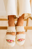 White sandals with ruffled details worn by a person sitting on a wooden bench.