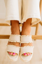 White sandals with ruffles worn by a person sitting on a wooden bench.
