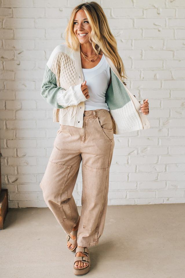Woman wearing a light jacket, white top, and beige pants against a white brick wall.